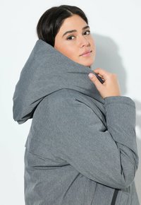 Chaqueta con capucha gris, fabricada con un tejido suave y texturizado, que presenta un corte holgado, una capucha prominente y un sutile detalle en las costuras.