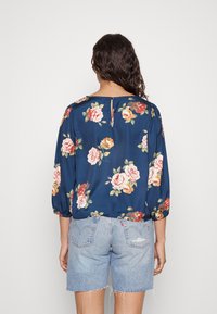 Blusa floral azul marinho com mangas três quartos, apresentando rosas rosas e amarelas. O design das costas inclui uma pequena abertura em forma de olho de fechadura na gola.