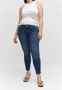 Woman wearing a white sleeveless top, blue skinny jeans, and black flip-flop sandals, standing against a plain background.