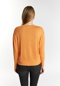 Oranger Strickpullover mit langen Ärmeln, Rundhalsausschnitt und lässiger Passform, kombiniert mit dunklen Jeans. Glatte Textur, minimale Details.