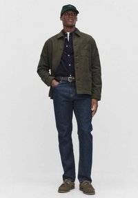 Grøn bomuldsjakke over en mørk skjorte, parret med blå jeans og brune sko. Outfittet har rene linjer og en struktureret pasform.