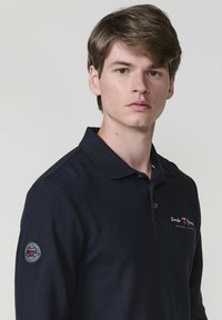 Sötétkék pólóing textúrált anyagból, gallérral, gombzárral és hímzett logóval a mellkason és az ujjan.