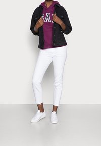 Černá džínová bunda, fialová mikina s bílým textem, bílé zkrácené kalhoty a bílé tenisky. Ležérní outfit s vrstvenými texturami a barvami.