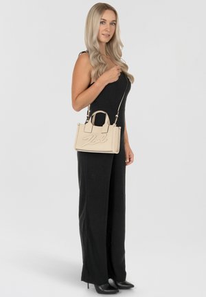 Beige Lederhandtasche mit geprägt Logo, strukturierter Form, Obergriffen und Schultergurt. Modell trägt einen schwarzen Jumpsuit und hohe Schuhe.
