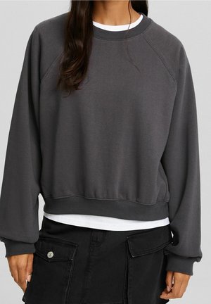Femme portant un sweat-shirt gris foncé à col rond sur une chemise blanche et une jupe cargo noire avec poches et boutons.