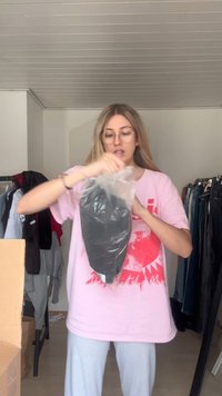 Sort plagg i plastinnpakning holdt av en person i en rosa skjorte, omgitt av klær på kleshengere og esker i et lyst innendørsområde.