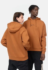 Sudadera con capucha marrón y un bolsillo delantero, con un logo bordado en negro. Corte holgado y puños de canalé, llevada por dos modelos.