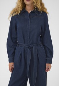 Frau trägt einen dunkelblauen Denim-Overall mit langen Ärmeln, einem Kragen und einem gebundenen Taillengürtel.