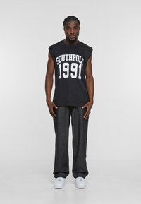 Schwarzes, ärmelloses T-Shirt mit weißem "SOUTHPOL 1991"-Text, kombiniert mit dunklen Jeans und weißen Sneakern, vor einem schlichten Hintergrund.