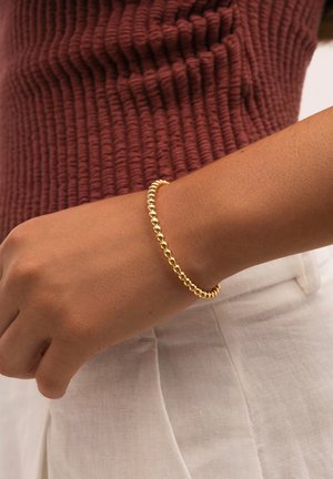Pulsera de cuentas doradas en una muñeca, con cuentas esféricas de igual tamaño y textura suave. Llevada con una blusa burdeos con relieve.