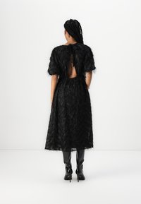 Robe en dentelle noire avec des manches courtes et bouffantes, une découpe au dos et une texture superposée avec un motif en zigzag. Portée avec des bottes noires montantes.