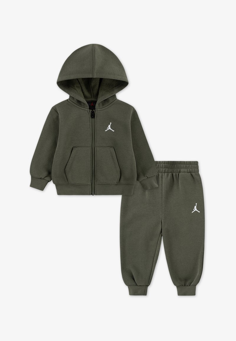 Φουτουριστικό σακάκι με φερμουάρ και κουκούλα σε ελιά πράσινο και παντελόνι που ταιριάζει, κατασκευασμένα από απαλό ύφασμα. Διαθέτουν λευκές λεπτομέρειες με λογότυπο και πλαϊνές τσέπες στο σακάκι.