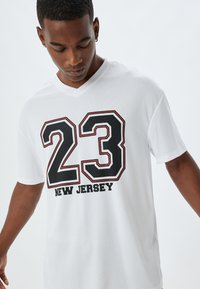 Biała koszulka t-shirt z czarną i czerwoną grafiką, na której widnieje numer "23" oraz napis "NEW JERSEY". Posiada dekolt w serek, krótkie rękawy oraz teksturowaną tkaninę.