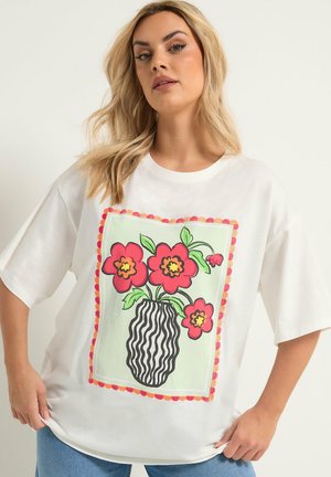 Blonde vrouw die een loszittend wit T-shirt draagt met een afbeelding van rode bloemen in een zwart-wit gestreepte vaas op een lichtgroene vierkante achtergrond.