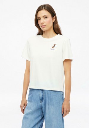Jonge vrouw met lang bruin haar die een wit T-shirt met korte mouwen draagt met een kleine hondenafbeelding en lichtblauwe wijde jeans.