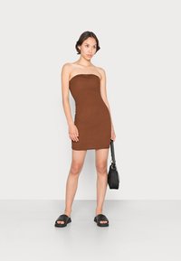 Vestido mini castanho, com costura canelada, sem alças e com silhueta ajustada. Combinado com sandálias de slide em couro preto e uma pequena bolsa preta.