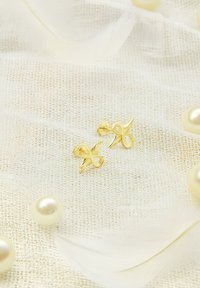 Goldene Steckerohrringe mit Unendlichkeit-Design, das eine glatte Textur und feine Details aufweist, vor einem sanften, hellen Hintergrund mit Perlen.