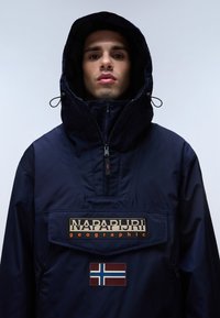 Tmavě modrá bunda s kapucí, na přední straně se zipem a velkou nášivkou loga; obsahuje odznak norské vlajky a texturované detaily látky.