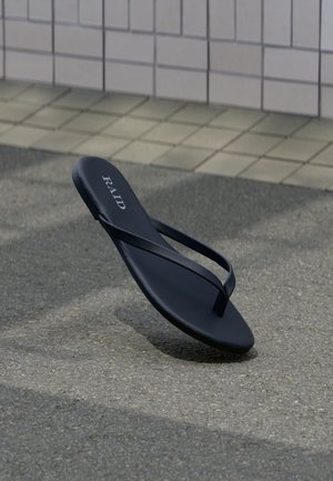 Sandale noire type tongs avec le logo "RAID" posée sur un pavement texturé près d'un trottoir carrelé en arrière-plan.