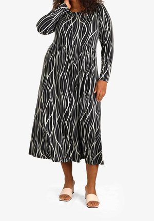 Kobieta ubrana w czarną sukienkę midi z długim rękawem, ozdobioną białymi abstrakcyjnymi falistymi liniami, w połączeniu z beżowymi sandałami na słupku z odkrytymi palcami.