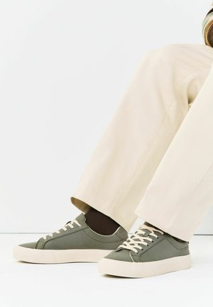 Vihreät matalat tennarit, joissa valkoiset pohjat ja nauhat, käytössä tummien sukkien ja vaaleiden beigen leveälahkeisten housujen kanssa valkoista taustaa vasten.