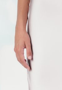 Uma mão esguia e bronzeada com unhas naturais repousa sobre um fundo de tecido liso e claro, destacando uma textura de pele clara.