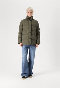 Olijfgroene pufferjack met een hoge kraag, vooraan knope sluitingen, gedragen over wijde blauwe jeans en zwarte sneakers met beige zolen.