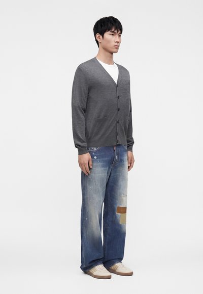Jeune homme portant un cardigan gris boutonné, une chemise blanche, un jean bleu ample avec des patchs et des baskets blanches, debout devant un fond uni.