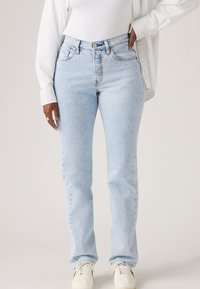 Άτομο που φοράει ανοιχτό μπλε τζιν παντελόνι με ίσια γραμμή και ψηλή μέση, λευκά αθλητικά παπούτσια, λευκό αμάνικο τοπ και λευκό πουκάμισο με μακριά μανίκια και κουμπιά, με το χέρι στην τσέπη.