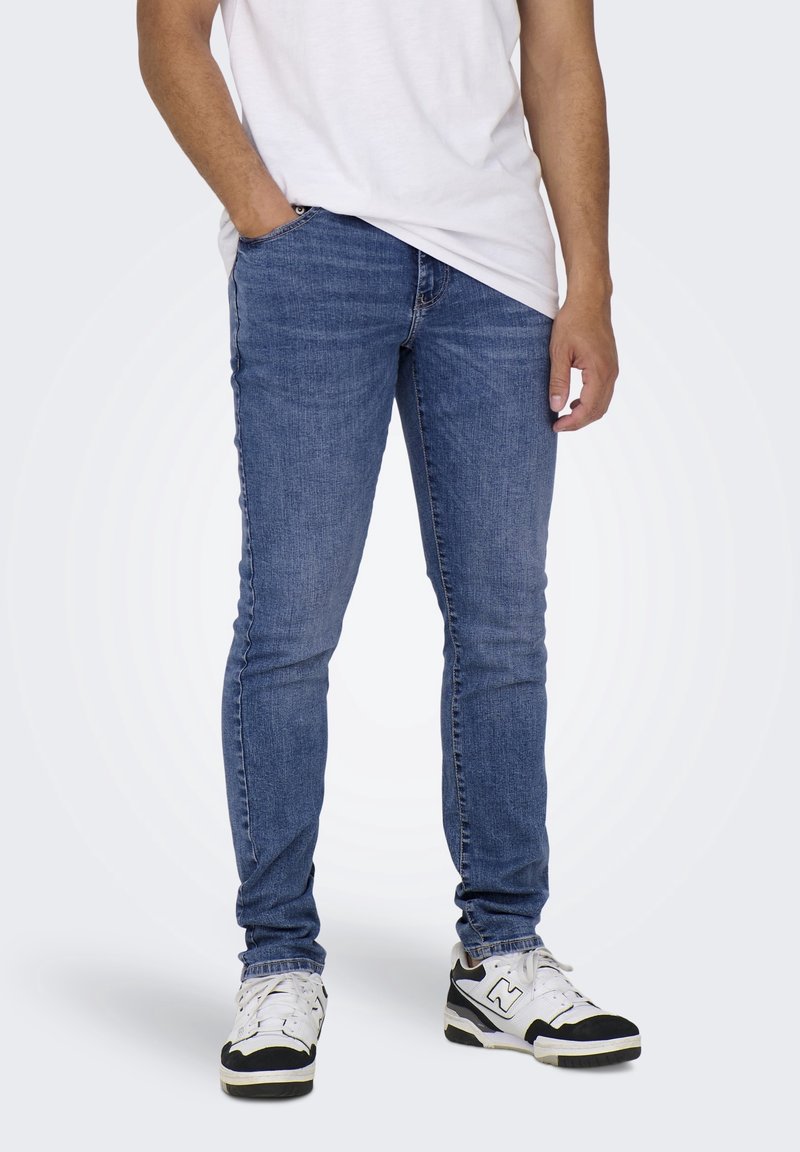 Hoikka siniset farkut, joissa on haalistunut pinta, yhdistettyinä yksinkertaiseen valkoiseen t-paitaan ja musta-valkoisiin tennareihin. Ei näkyviä bränditietoja.