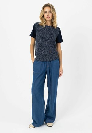 Lyshåret kvinde iført navy kortærmet top med tekstureret ærmeløs overlay, vide blå jeans og beige mokassinsko, stående med hænderne i lommerne.