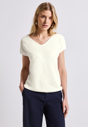 Haut blanc texturé à col en V avec des manches courtes, arborant un motif subtil, associé à un pantalon noir marine.