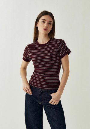 Jonge vrouw die een nauwsluitend, bordeauxrood-wit gestreept shirt met korte mouwen en donkerblauwe spijkerbroek draagt, staand met één hand in haar zak.