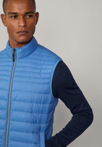 Ljusblå vadderad väst med hög krage, med horisontell kviltad textur och en framtida dragkedja. Bärs över en marinblå långärmad skjorta.