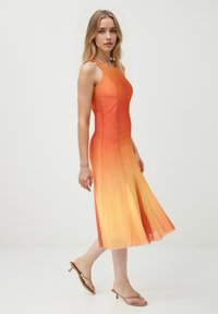 Robe dégradée orange à jaune avec des bretelles spaghetti, un corsage ajusté, une jupe évasée et une texture transparente. Associée à des sandales minimalistes.