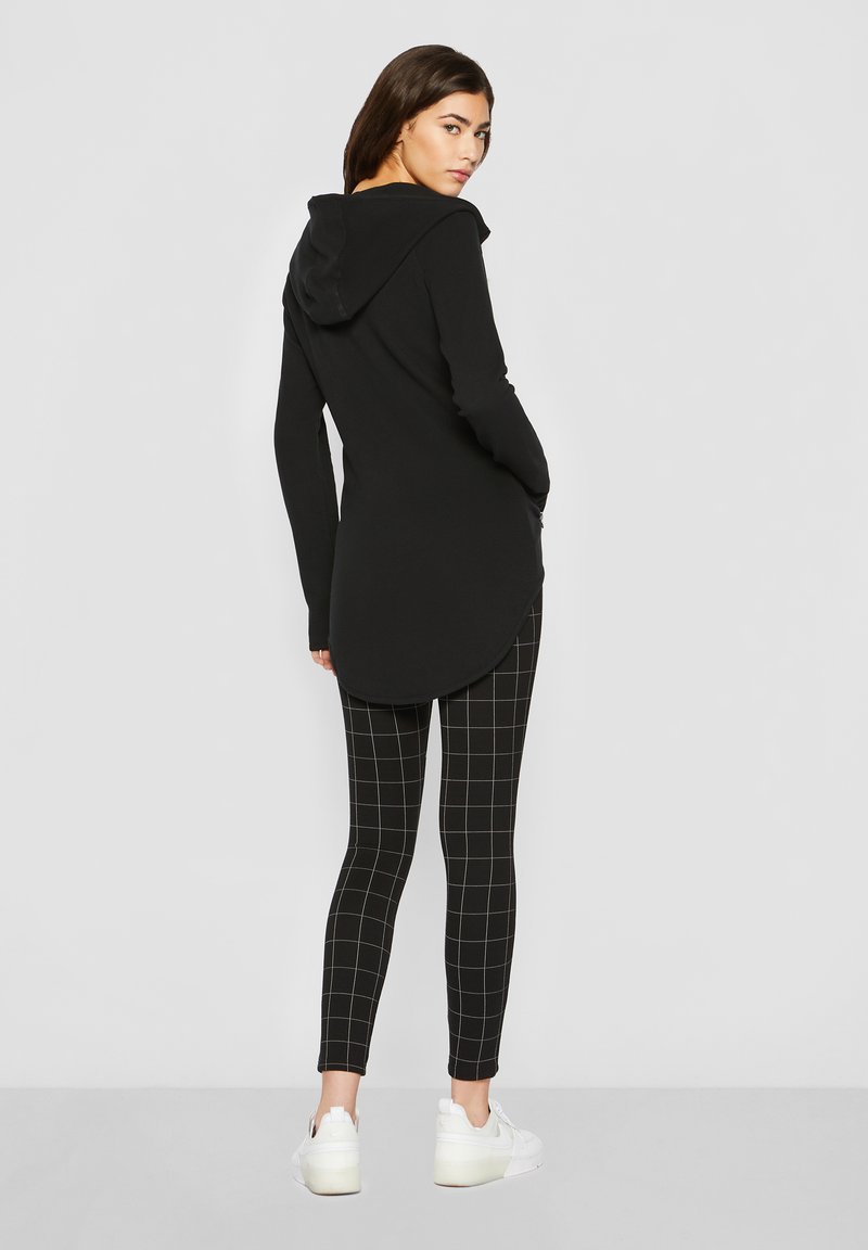 Femme en haut noir long à capuche, leggings noirs avec un motif à grille blanche, et baskets blanches, debout de dos, tête tournée.