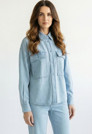 Frau mit langem, welligem Haar, die ein hellblaues Jeanshemd mit geknöpften Taschen und passende Jeanshose vor einfachem Hintergrund trägt.