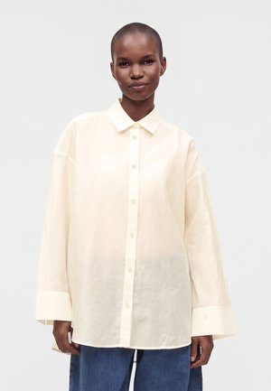 Persona que lleva una camisa de botones suelta de color crema con mangas largas y pantalones azules sobre un fondo liso y claro.