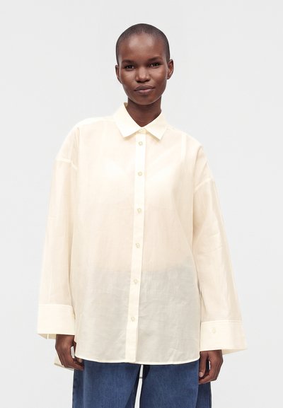 Personne portant une chemise ample crème à boutons avec manches longues et un pantalon bleu sur un fond clair uni.