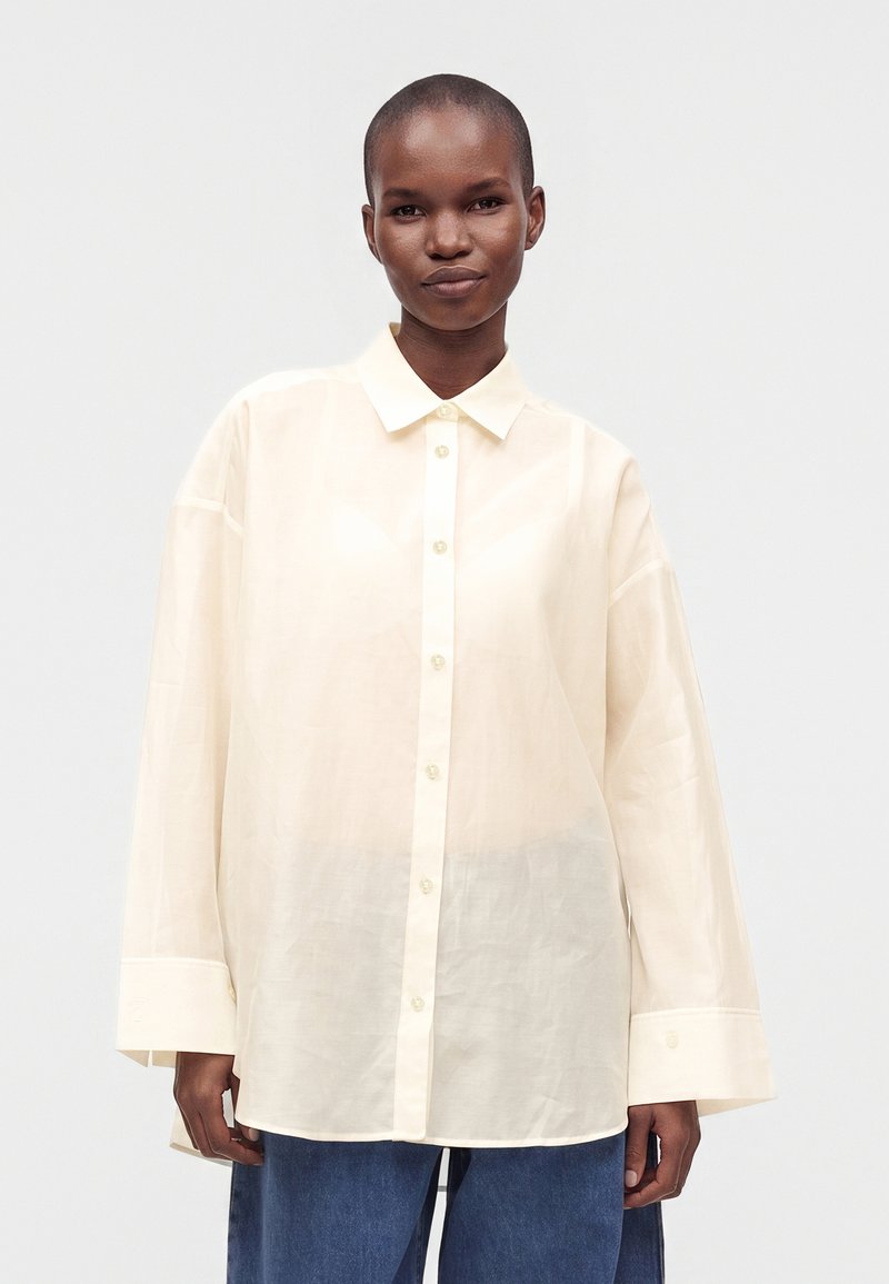 Personne portant une chemise ample crème à boutons avec manches longues et un pantalon bleu sur un fond clair uni.