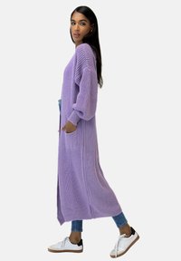 Lång, öppen cardigan i mjuk lavendelstickning. Har breda ärmar, två framfickor och är kombinerad med blå jeans och sneakers.