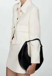 Sac à main en cuir noir à la forme arrondie, texture lisse et poignée fine, tenu à côté d'une veste blanche texturée ornée de boutons dorés.