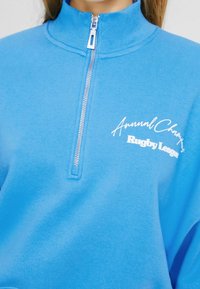Sudadera azul con cremallera en tela suave, con cuello alto y texto blanco "Annual Champions Rugby League" en el frente. Detalle de cremallera plateada.
