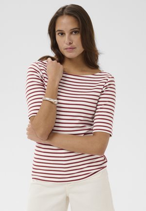 Femme aux longs cheveux bruns portant une chemise blanche à rayures rouges et un pantalon crème, posant sa main sur son épaule opposée.