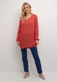 Czerwony tunik o długim rękawie i dekolcie w kształcie litery V, zestawiony z ciemnoniebieskimi spodniami. Tunik ma prosty krój, gładką fakturę i minimalne wykończenia.