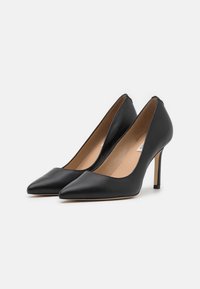 Chaussures à talons aiguilles en cuir noir avec un design à bout pointu, des bords arrondis élégants et un talon dénudé, présentant une semelle intérieure beige.