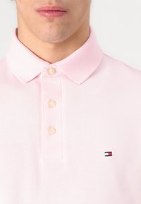 Moški nosi svetlo rožnato polo majico s tremi gumbi in majhnim rdeče-belo-modrim logotipom na prsih.