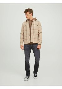 Károvaná bunda v béžových a hnedých odtieňoch s čiernymi gombíkmi, prehodená cez hnedú mikinu s kapucňou. Súčasťou outfitu sú tmavé džínsy a čierne tenisky.