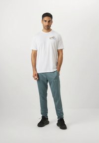 Valge lühikeste varrukatega T-särk logoga, kombineeritud türkiissiniste spordipükste ja mustade tossudega. Modell seisab ühevärvilise tausta ees.