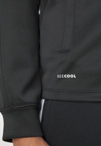 Sort jakkeærme og lommeområde med hvid tekst "BEE COOL" nær taljen, båret over matchende sorte bukser med en hvid stribe.